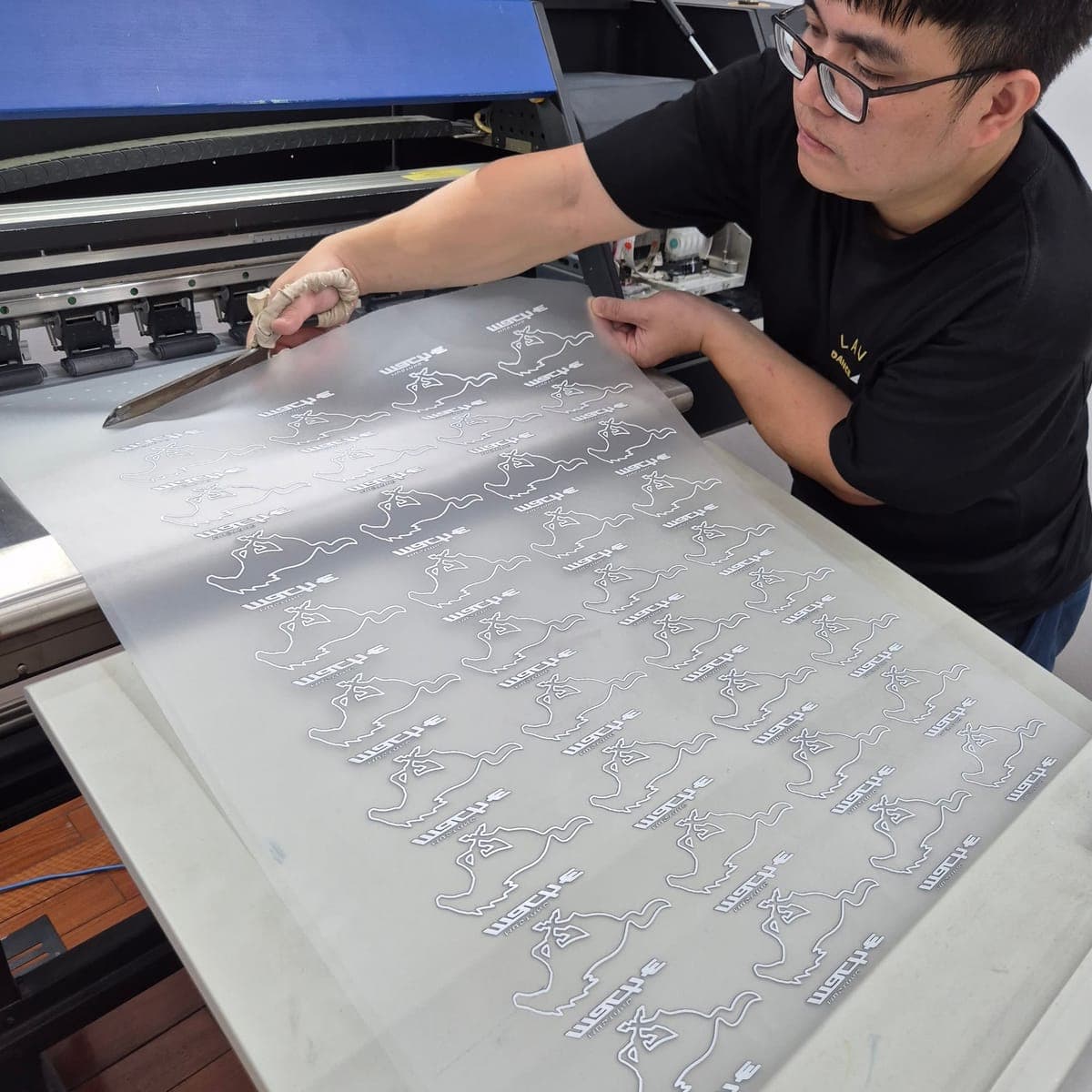 Thợ bóc màng film DTF vừa in, logo lặp lại thành nhiều hình, cạnh máy in DTF khổ lớn tại xưởng Startee
